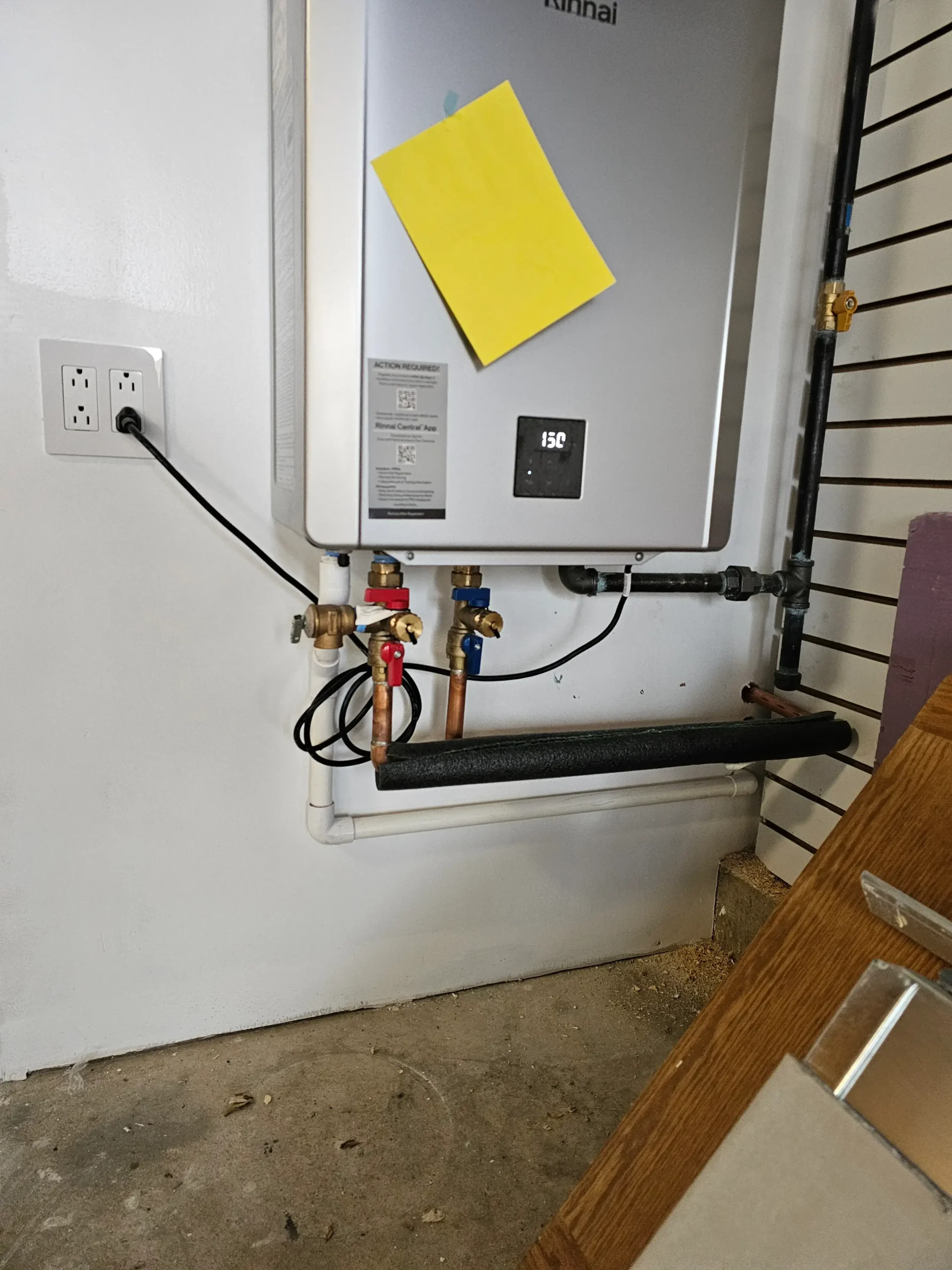 Tankless water heater installation with exposed plumbing lines, shutoff valves, and electrical outlet mounted on wall in utility space.