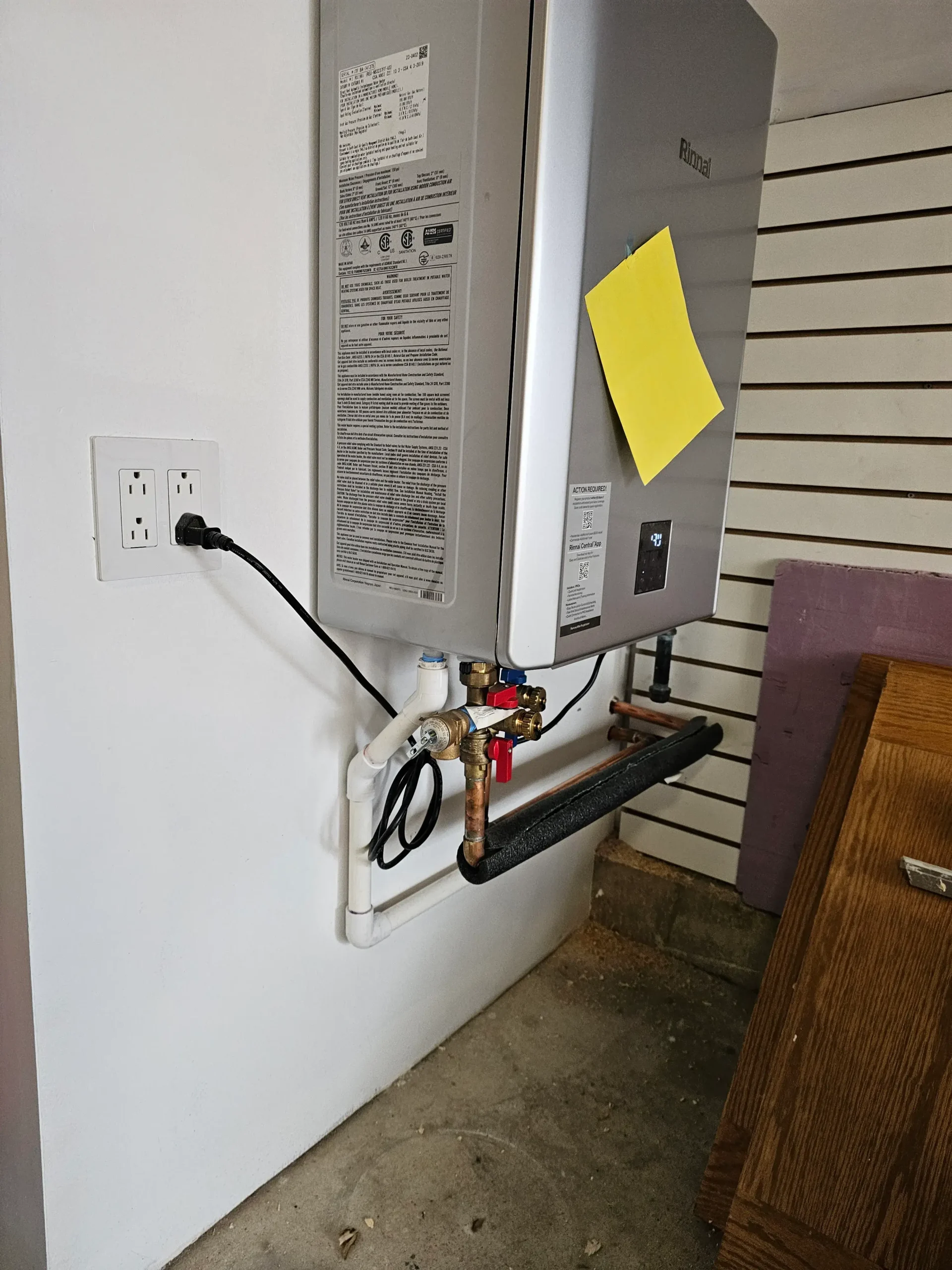 Modern tankless water heater mounted on wall with copper piping, water valves, and drainage setup in a residential utility area.
