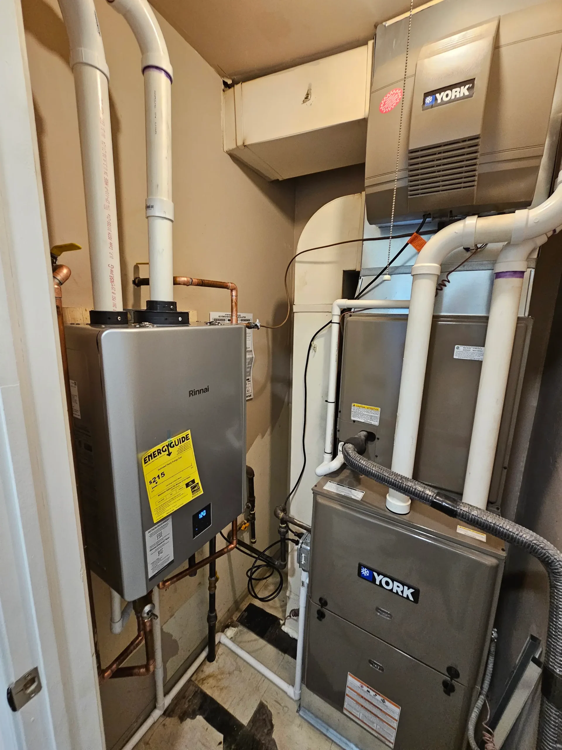 Tankless water heater installed next to furnace system with PVC venting, copper piping, and gas line connections in a residential utility room.