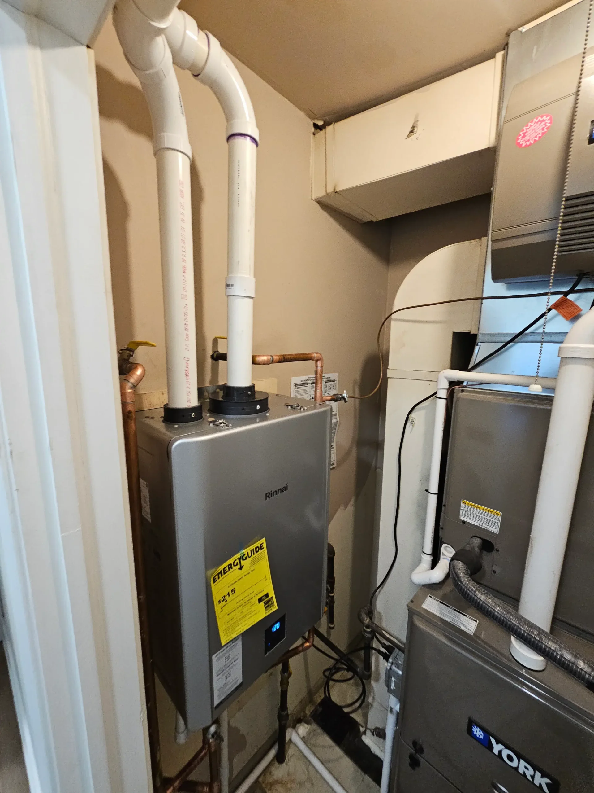 tankless water heater with dual vent pipes and copper plumbing lines connected beside HVAC furnace in a compact mechanical space